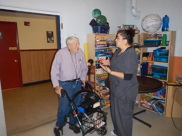 An elderly man with a walker is talking to a woman in gray scrubs in a room with shelves filled with board games and other recreational items.