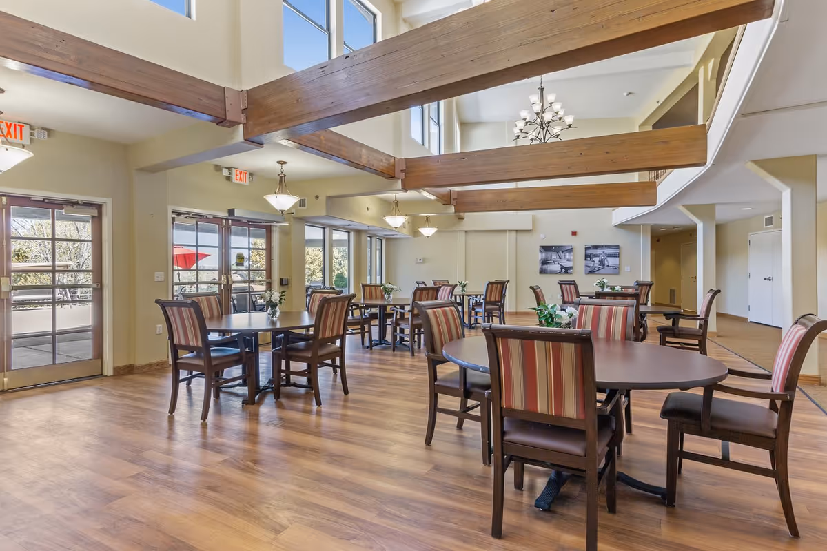 A spacious dining room with multiple round tables and striped cushioned chairs. The room features large windows and glass doors letting in natural light, wooden ceiling beams, and hanging light fixtures. There are small flower arrangements on each table and framed pictures on the far wall.