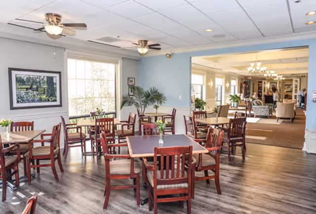 A bright dining room with several wooden tables and chairs arranged neatly. The room has large windows letting in natural light, ceiling fans, and light blue walls. There are small vases with flowers on the tables and a view into an adjacent lounge area with more seating and plants.