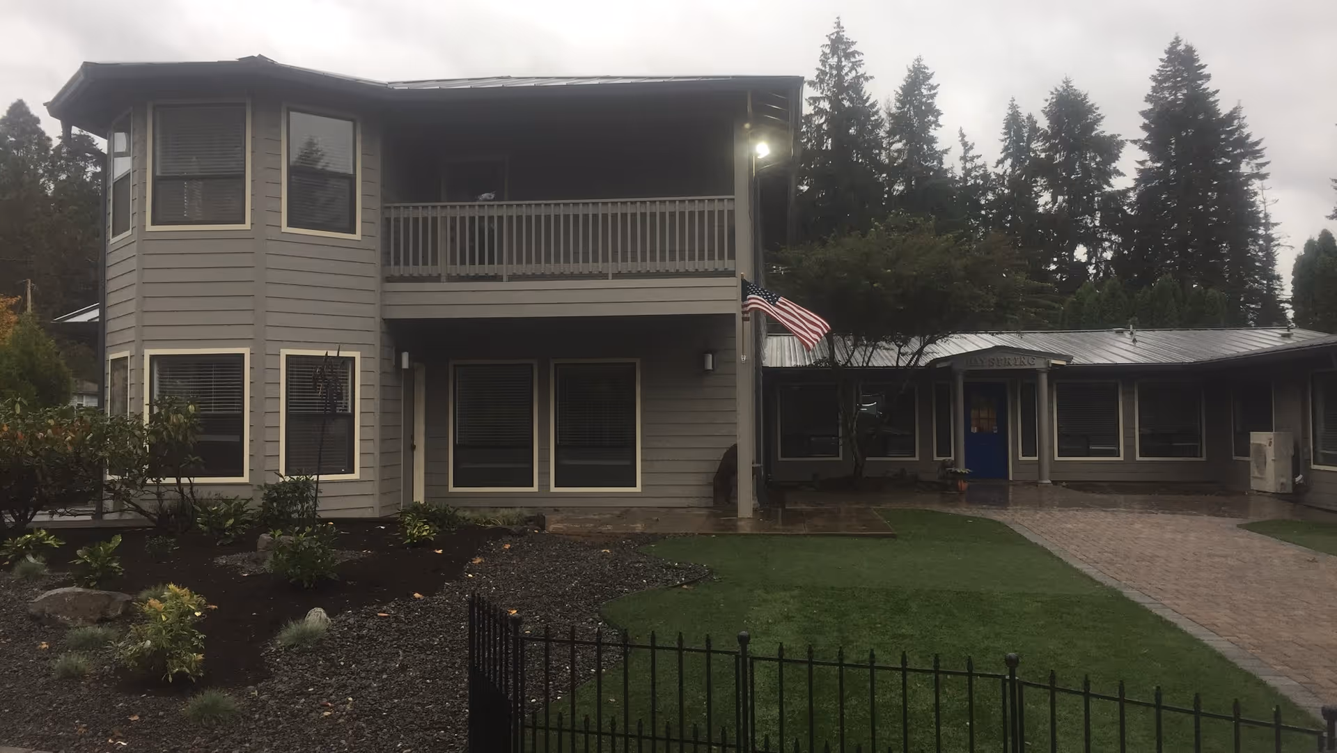 Exterior view of a two-story building with gray siding and multiple windows, featuring a balcony on the upper floor. There is a paved walkway leading to a blue door under a small covered entrance. The area in front includes a lawn, some bushes, and trees, with an American flag mounted on a pole near the entrance. The sky is overcast.