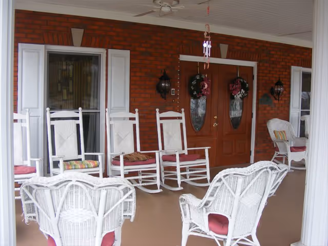 Covered porch area with white rocking chairs and wicker chairs with red cushions arranged around a brown floor. The porch has a brick wall with two wooden doors decorated with floral wreaths and two wall-mounted lantern lights on either side of the doors.