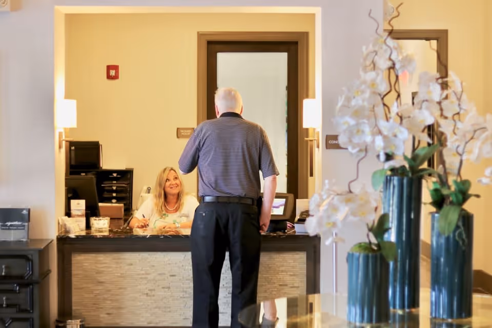 An elderly man standing at a reception desk talking to a smiling receptionist in a well-lit lobby area with decorative white orchids in blue vases in the foreground.