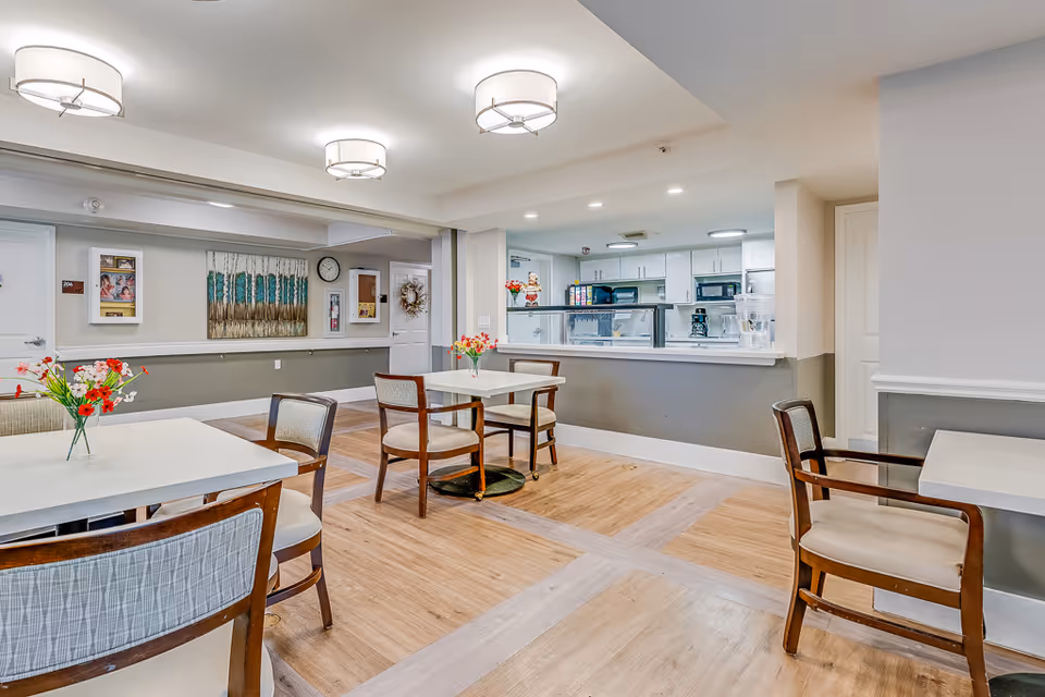 A bright and clean dining area in a senior living facility with several tables and chairs arranged neatly. The room features light wood flooring, modern ceiling lights, and a serving counter with a kitchen visible behind it. There are decorative flowers on the tables and artwork on the walls.