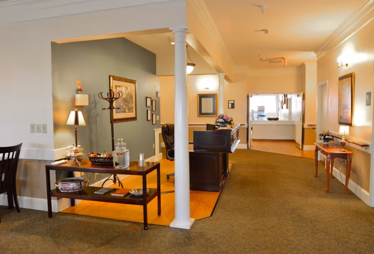 Interior view of a senior living facility reception area with a wooden reception desk, a coat rack, framed artwork on the walls, a small table with a lamp and flowers, and a hallway leading to another room with a counter and window.