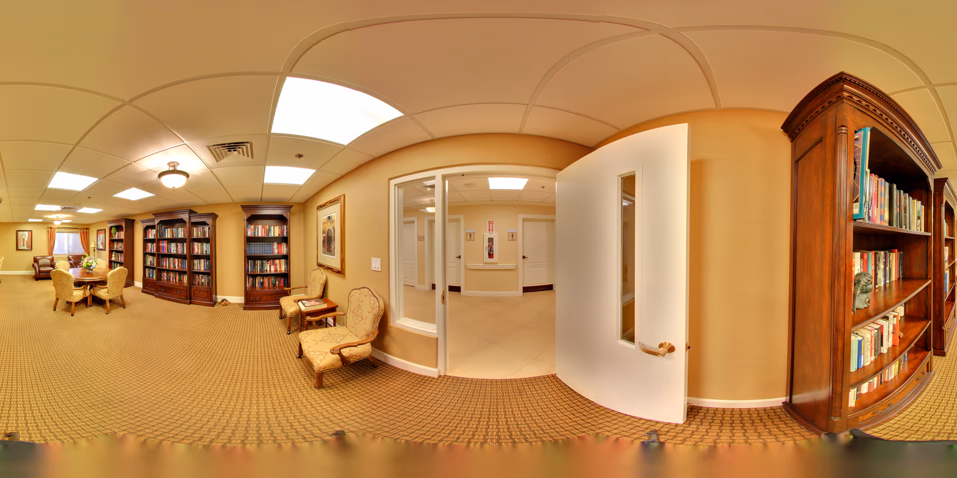 A spacious and well-lit interior common area in a senior living facility featuring multiple wooden bookshelves filled with books, a round table with four upholstered chairs, two additional upholstered chairs against the wall, framed artwork, and a white door leading to a hallway with beige walls and tiled flooring.