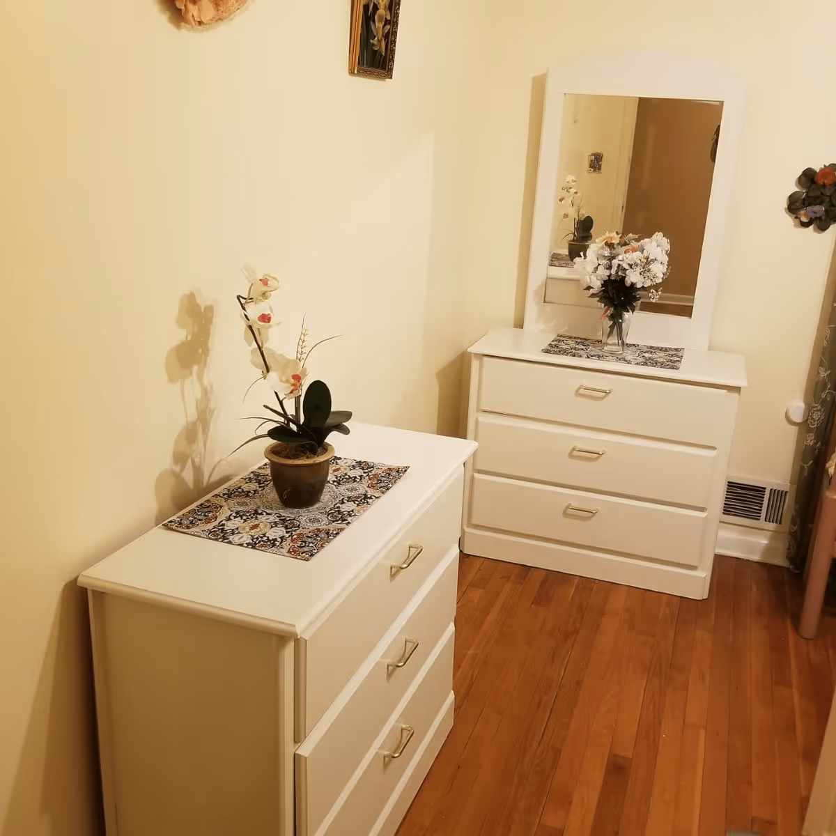 Narrow bedroom area with two white dressers, a mirror, and potted flowers on a hardwood floor.