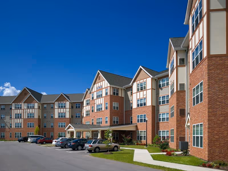 Exterior view of a multi-story senior living facility building with brick and beige siding under a clear blue sky. Several cars are parked in front along a paved driveway with landscaped greenery and a sidewalk leading to the entrance.
