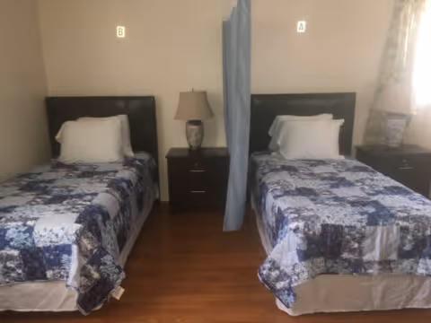 A bedroom with two single beds separated by a nightstand with a lamp. Each bed has a dark headboard and is covered with a blue and white patchwork quilt. A curtain hangs between the beds. There is a window with curtains on the right side of the room.