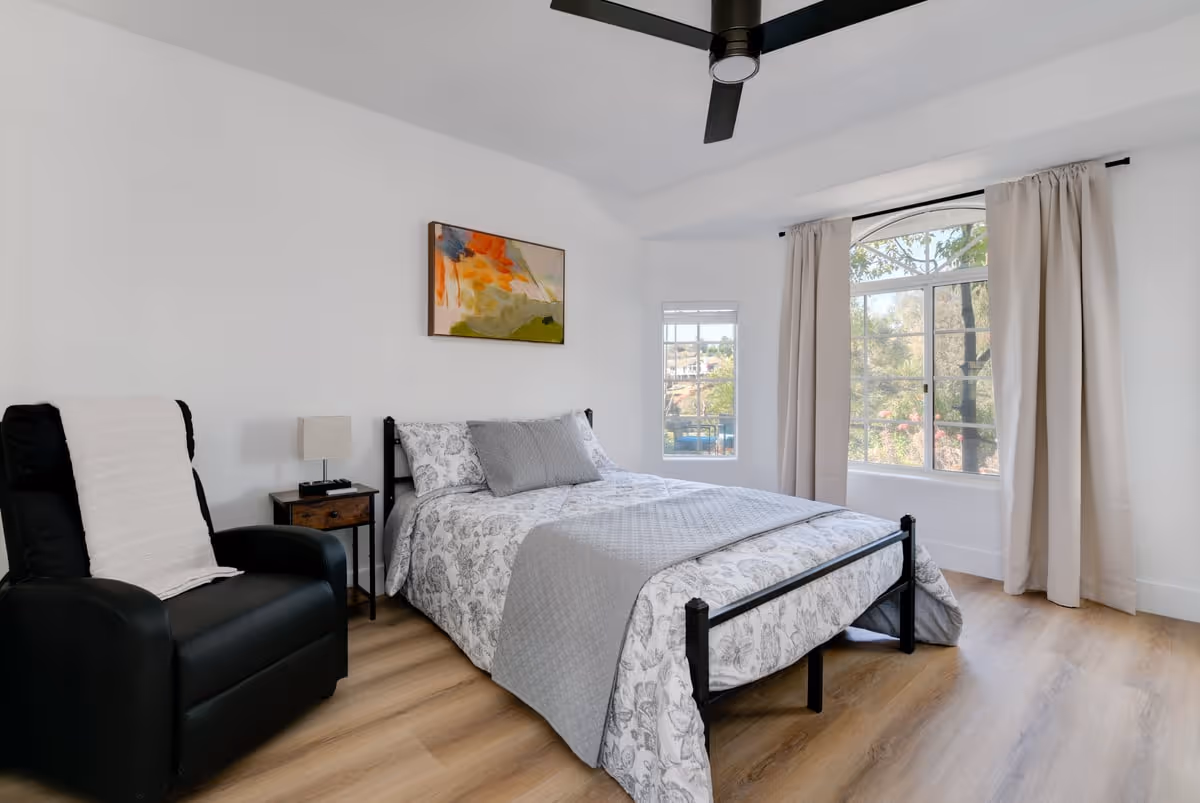 A bright bedroom with a large window covered by beige curtains, a bed with patterned bedding and gray pillows, a black recliner chair with a white throw, a small wooden nightstand with a lamp, and a ceiling fan above. There is a colorful abstract painting on the white wall.
