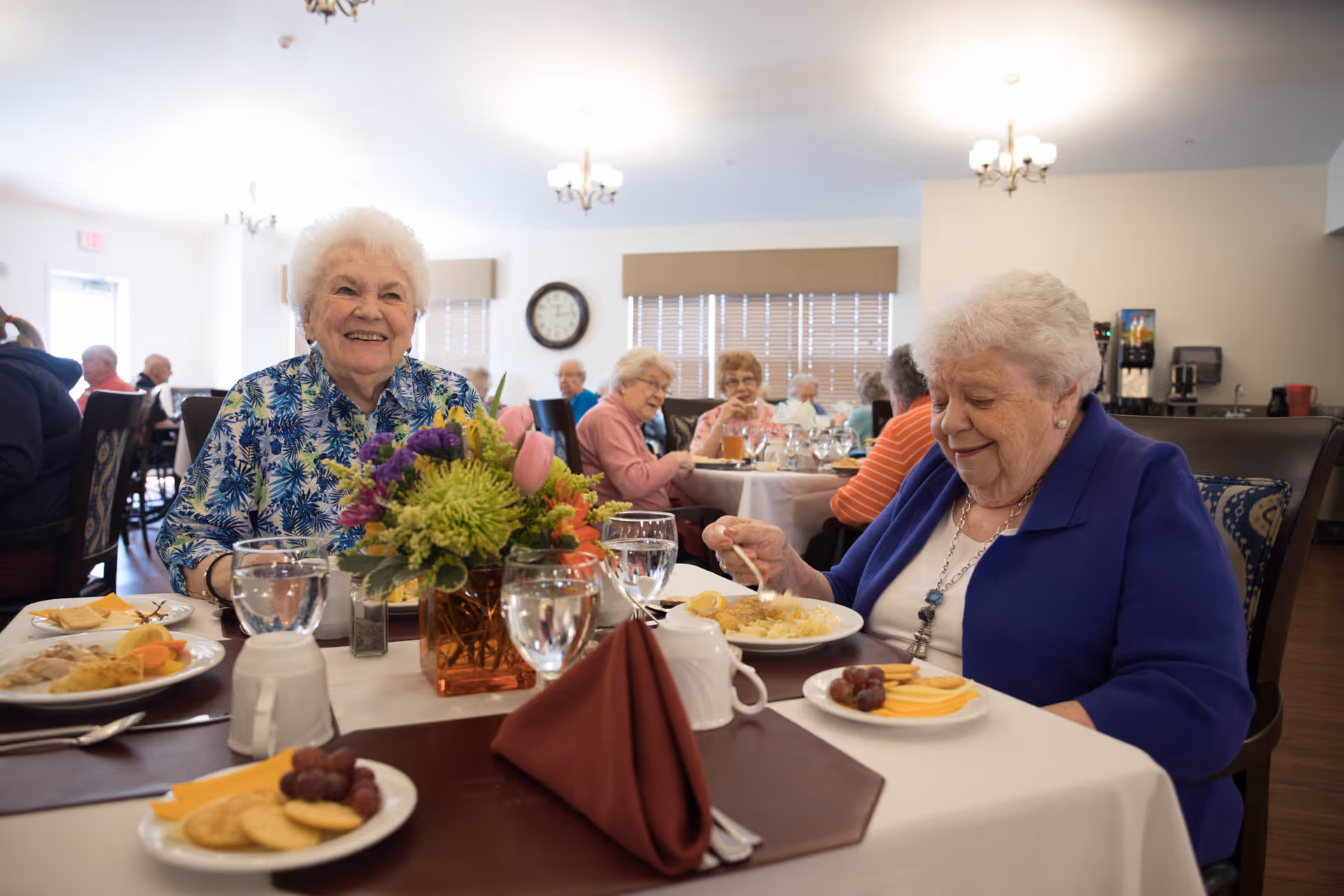 Two elderly women sitting at a dining table in a well-lit room, enjoying a meal together. The table is set with plates of food, glasses of water, and a centerpiece with colorful flowers. Other elderly people are seated at tables in the background, and the room has a warm, welcoming atmosphere with chandeliers and large windows with blinds.