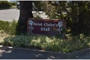 A small brown wooden sign with white text reading 'Saint Claire's 6248' placed among green bushes and trees near a paved area.