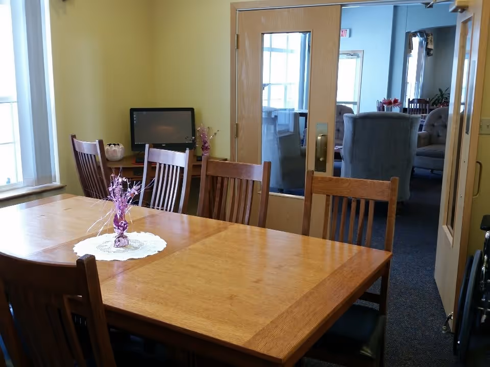 A wooden dining table with six chairs around it in a room with yellow walls. A small decorative centerpiece with purple flowers is on the table. In the background, there is a computer on a desk and a glass door leading to another room with upholstered chairs and a table.