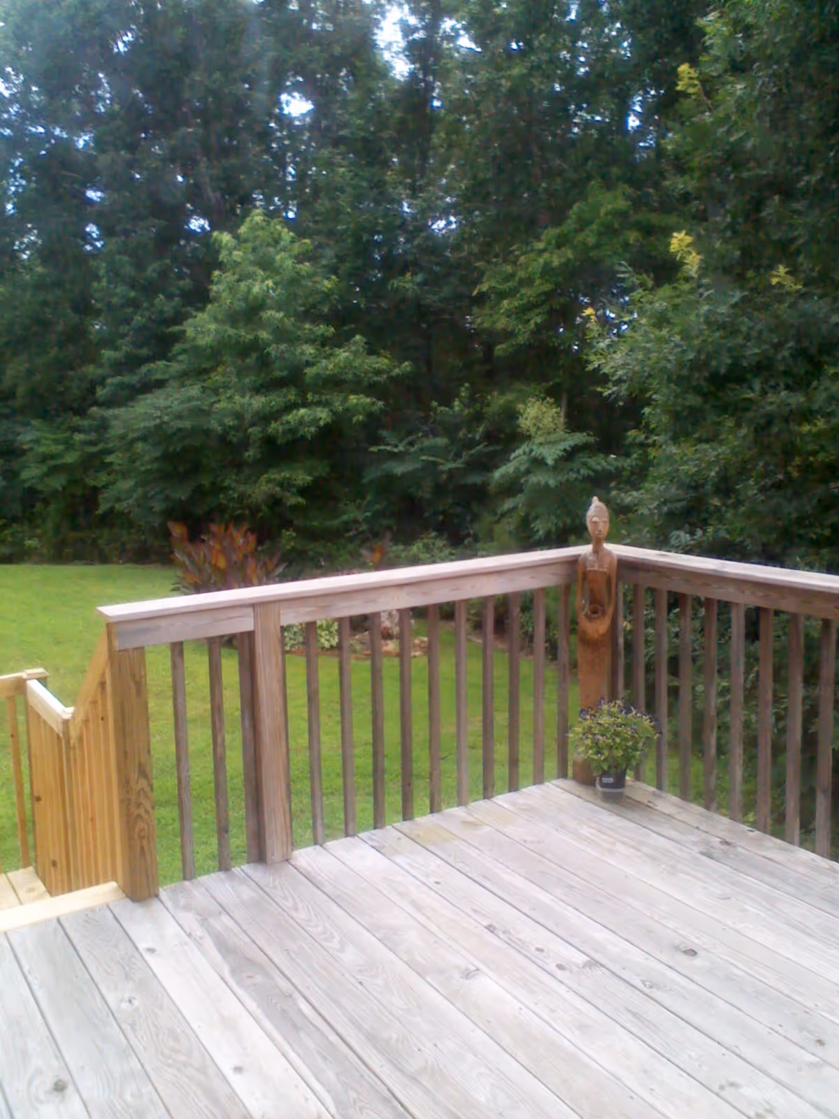 Wooden deck with railing overlooking a green grassy yard and dense trees in the background. A small potted plant and a wooden carved statue are placed on the deck near the corner of the railing.
