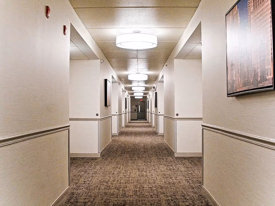 A long, well-lit hallway in a supportive living facility with beige walls, carpeted floor, and ceiling lights. The hallway has several doorways on both sides and framed artwork on the walls.