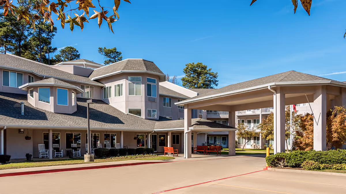 Exterior view of Asher Point Independent Living of Texarkana showing a multi-story building with large windows, a covered entrance with pillars, outdoor seating including benches and rocking chairs, and landscaped greenery under a clear blue sky.