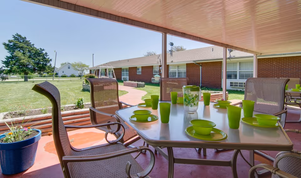 Covered outdoor patio area with a table set for six with green plates, bowls, cups, and a pitcher. Surrounding the patio is a grassy yard with a brick building and a swing set in the background under a clear blue sky.