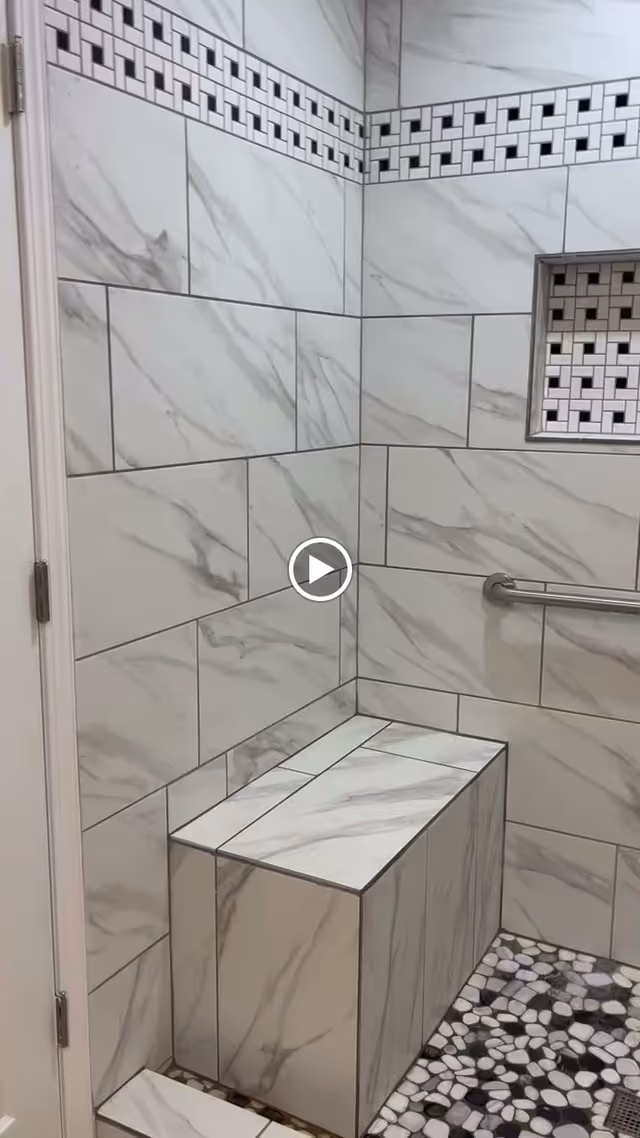 A tiled shower area with marble-patterned wall tiles and a built-in bench. The shower features a decorative tile border near the ceiling and a recessed niche with matching decorative tiles. There is a metal grab bar mounted on the wall and a pebble-style tiled floor with a drain.