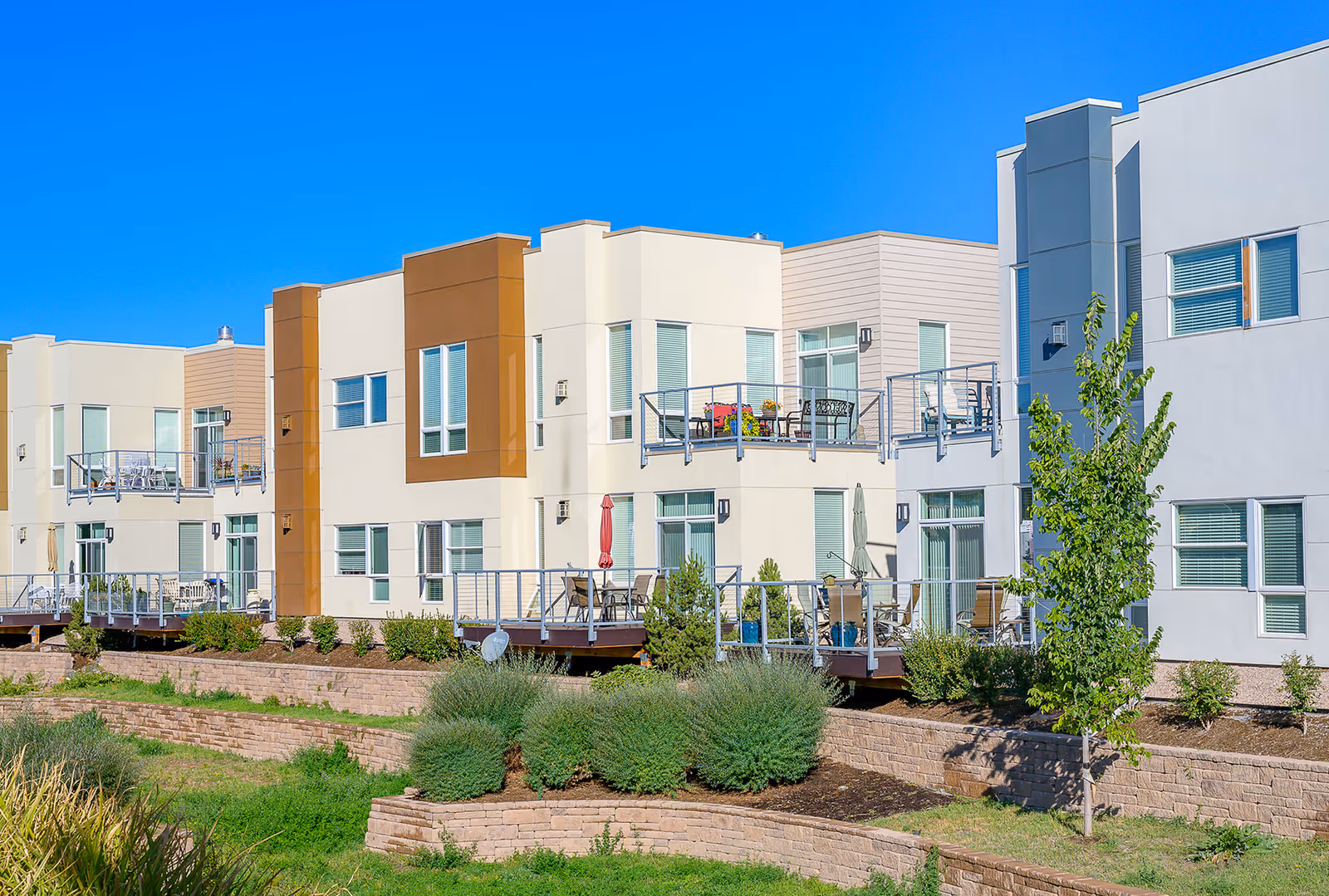 Exterior view of a modern senior living facility with multiple two-story units featuring balconies and patios. The building has a clean, contemporary design with beige, white, and brown accents. There are landscaped areas with shrubs, small trees, and grass in front of the building under a clear blue sky.
