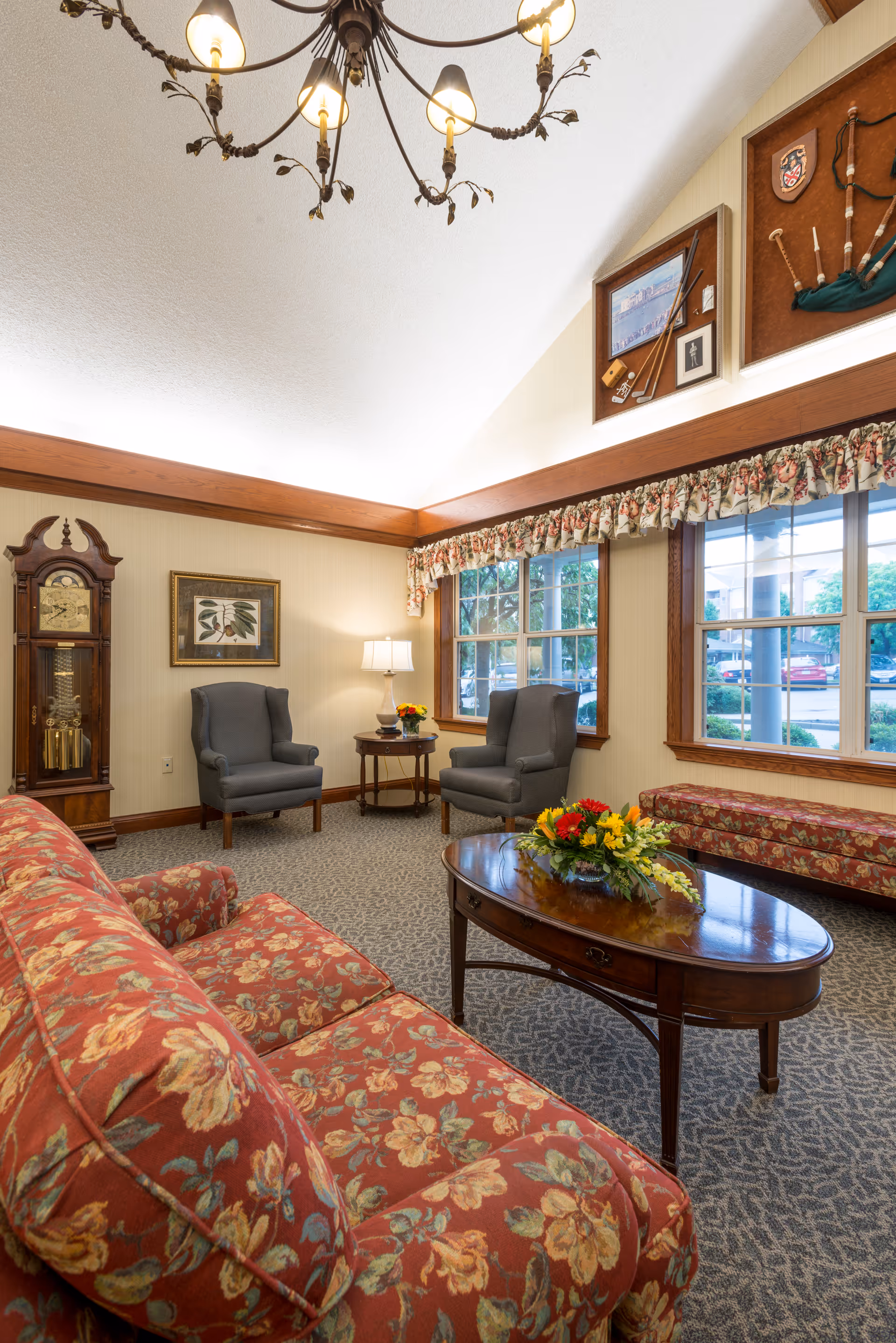 A cozy living room area with floral patterned sofa, two gray armchairs, a wooden coffee table with a flower arrangement, a grandfather clock, framed artwork on the wall, and large windows with floral valances letting in natural light.