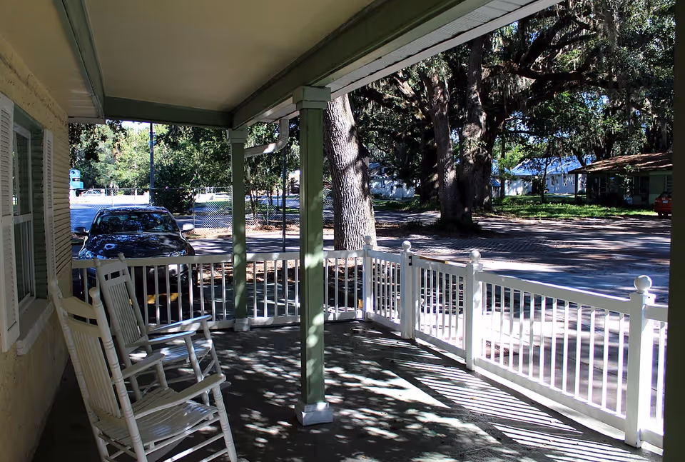 A covered porch area with two white rocking chairs and white railing. The porch overlooks a street with large trees and some parked cars in the background.