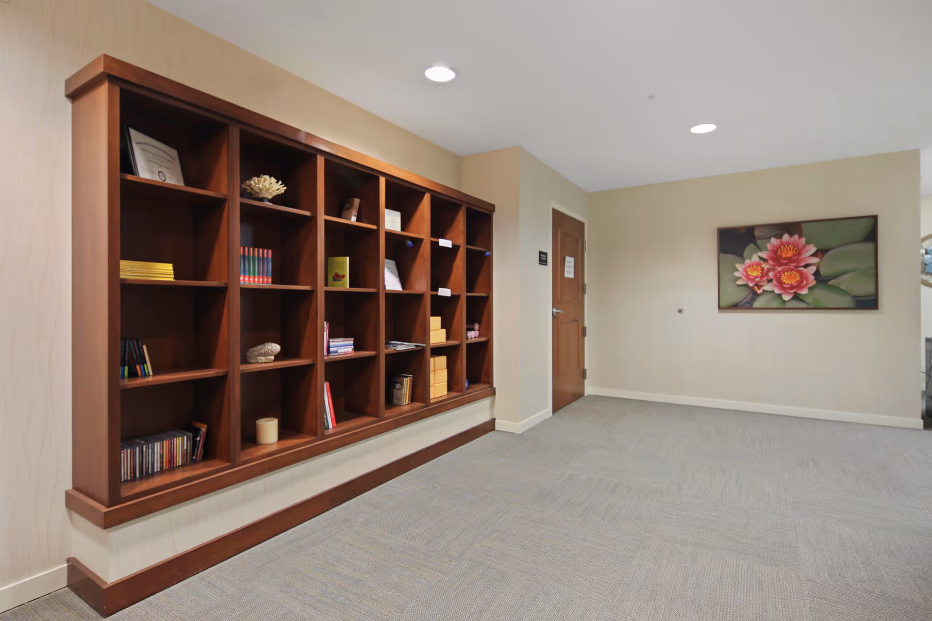 Interior hallway with a large wooden wall-mounted bookshelf containing books, decorative items, and boxes. There is a closed wooden door at the end of the hallway and a framed painting of pink water lilies on the wall to the right.