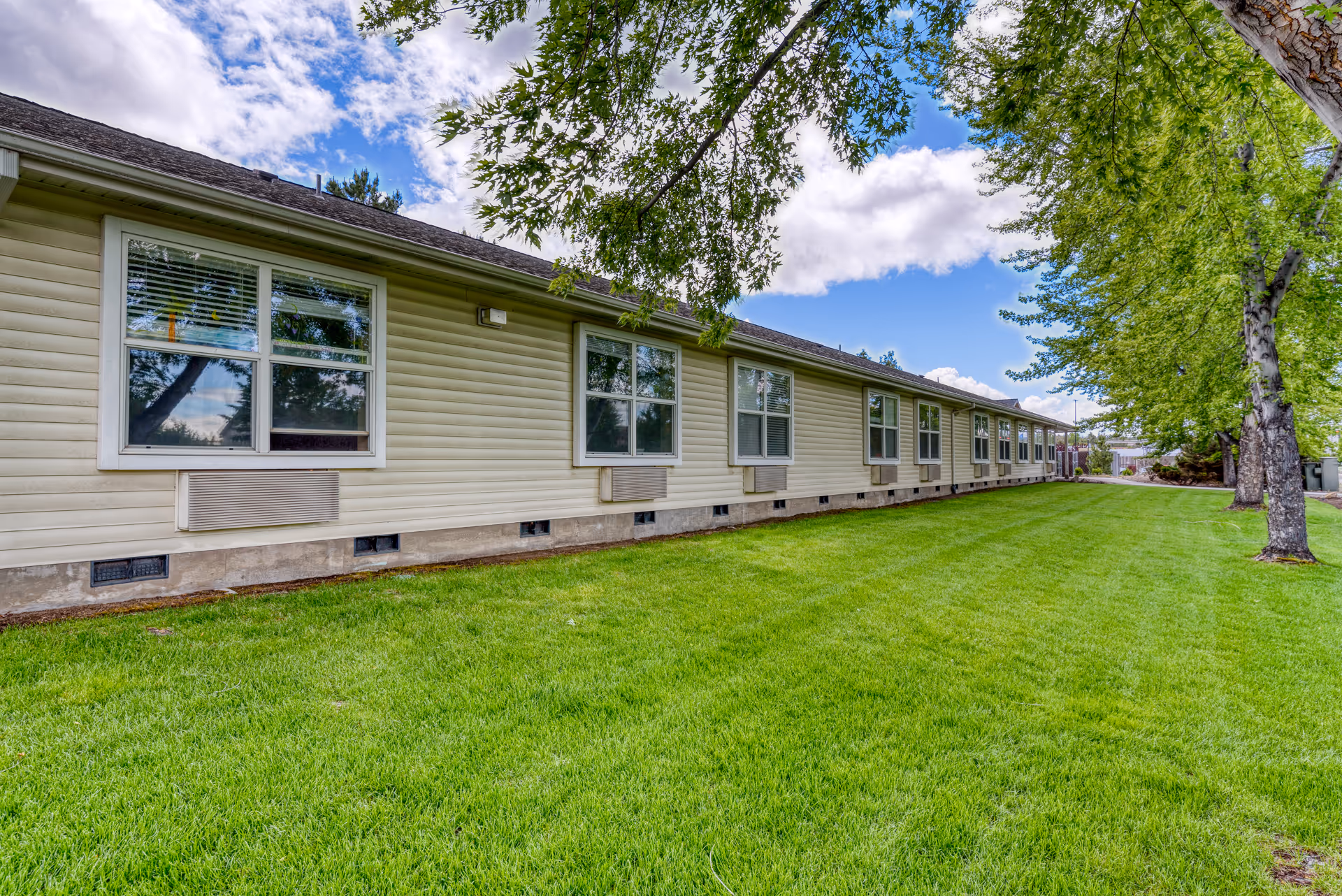 Exterior side view of a single-story building with beige siding and multiple windows, surrounded by a well-maintained green lawn and trees under a partly cloudy blue sky.