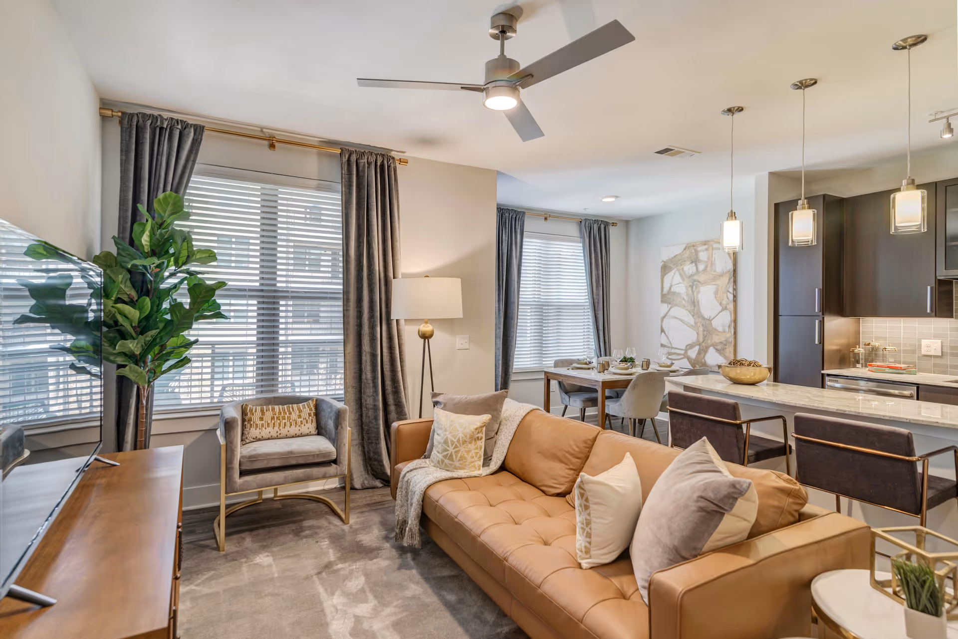 A modern living room with a tan leather sofa adorned with multiple pillows, a gray armchair, a floor lamp, and a large flat-screen TV on a wooden stand. The room has large windows with gray curtains and a ceiling fan with lights. In the background, there is a dining area with a table set for four and a kitchen with a marble countertop and three pendant lights hanging above it.