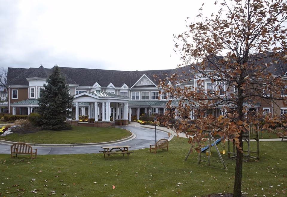 Exterior view of a senior living facility building with a covered entrance, surrounded by a circular driveway, green lawn, benches, a picnic table, a tree with brown leaves, and a small playground with swings under an overcast sky.