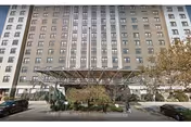 Front exterior view of a large multi-story residential building with numerous windows. There is a covered entrance with a metal canopy and some greenery including trees and bushes in front. Cars are parked along the street on both sides of the entrance.