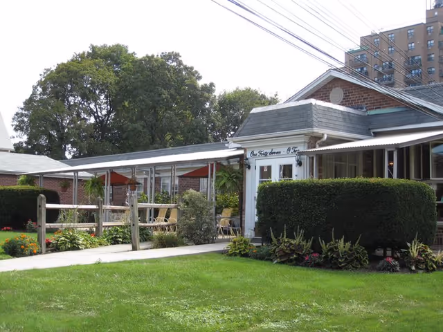Exterior view of a single-story brick building with a covered walkway and outdoor seating area with tables and chairs. There is a well-maintained lawn and landscaped bushes and flowers in the foreground. Trees and a taller building are visible in the background.