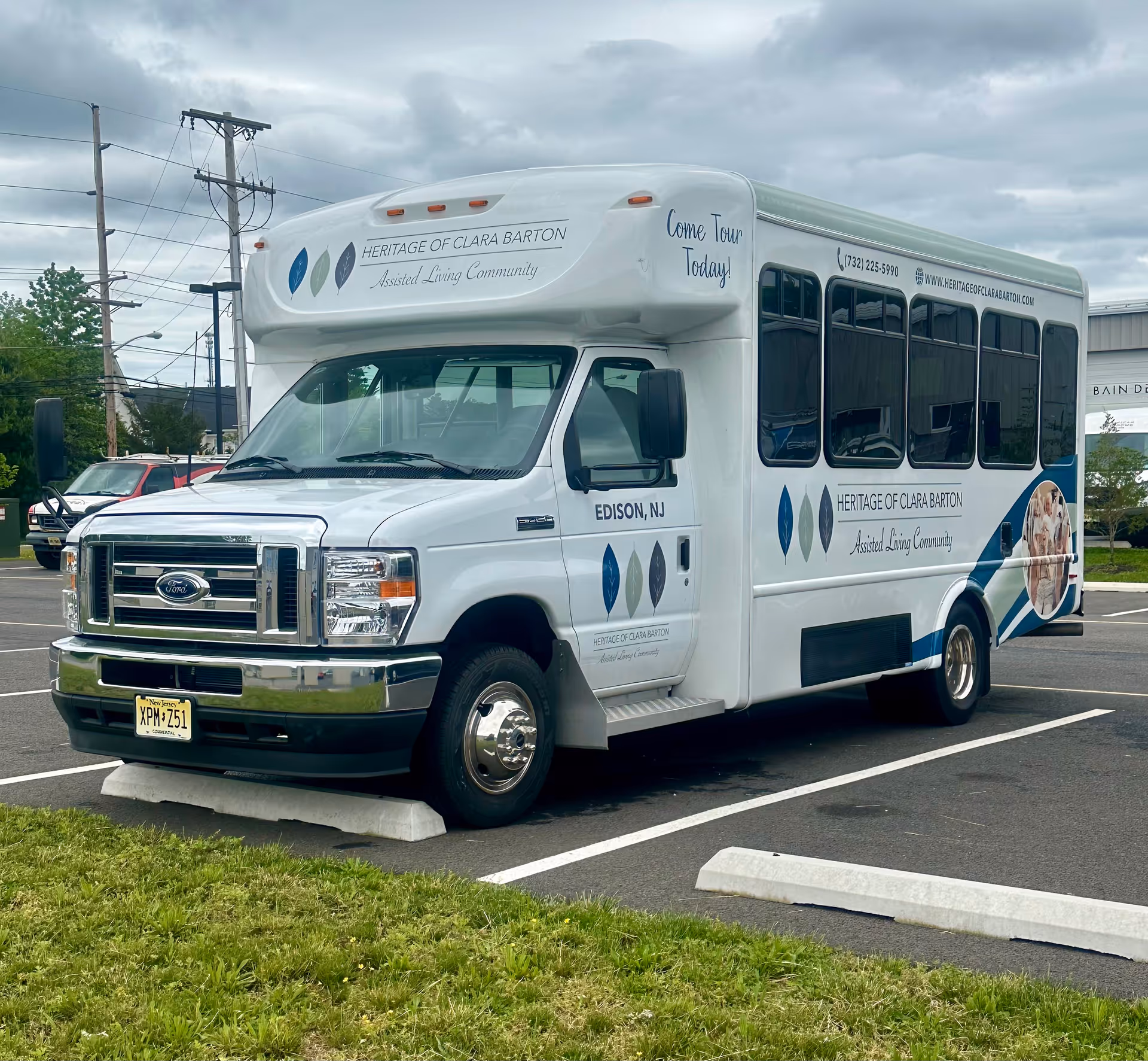 A white shuttle bus parked in a parking lot with signage for Heritage of Clara Barton Assisted Living Community. The bus has the text 'Come Tour Today!' and contact information including a phone number and website. The location is Edison, NJ.