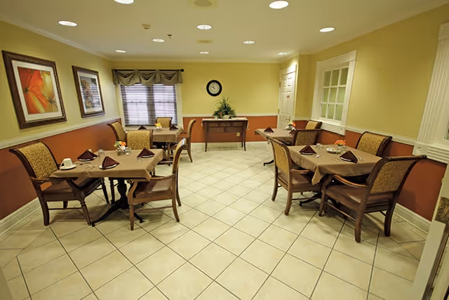 A dining room with four tables, each set with brown tablecloths, folded napkins, cups, and small flower arrangements. The room has beige tiled floors, yellow and brown walls, framed artwork, a window with blinds and a valance, a clock on the wall, and a small console table with a plant.