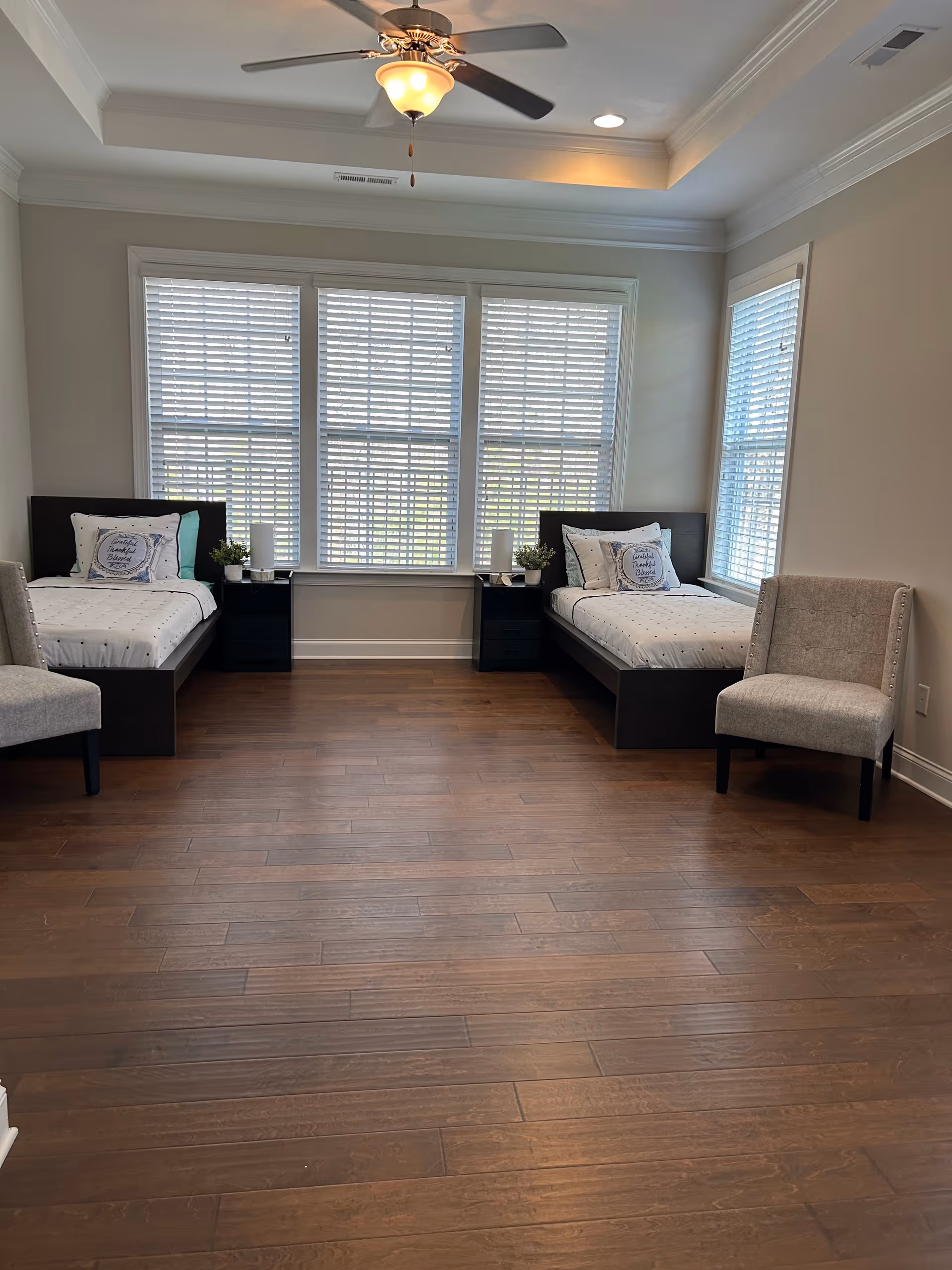 A bright bedroom with two single beds placed against opposite walls, each with white bedding and decorative pillows. Between the beds are two black nightstands with small plants and candles. Two beige upholstered chairs are positioned near the beds. The room has large windows with white blinds, a ceiling fan with a light fixture, and wooden flooring.
