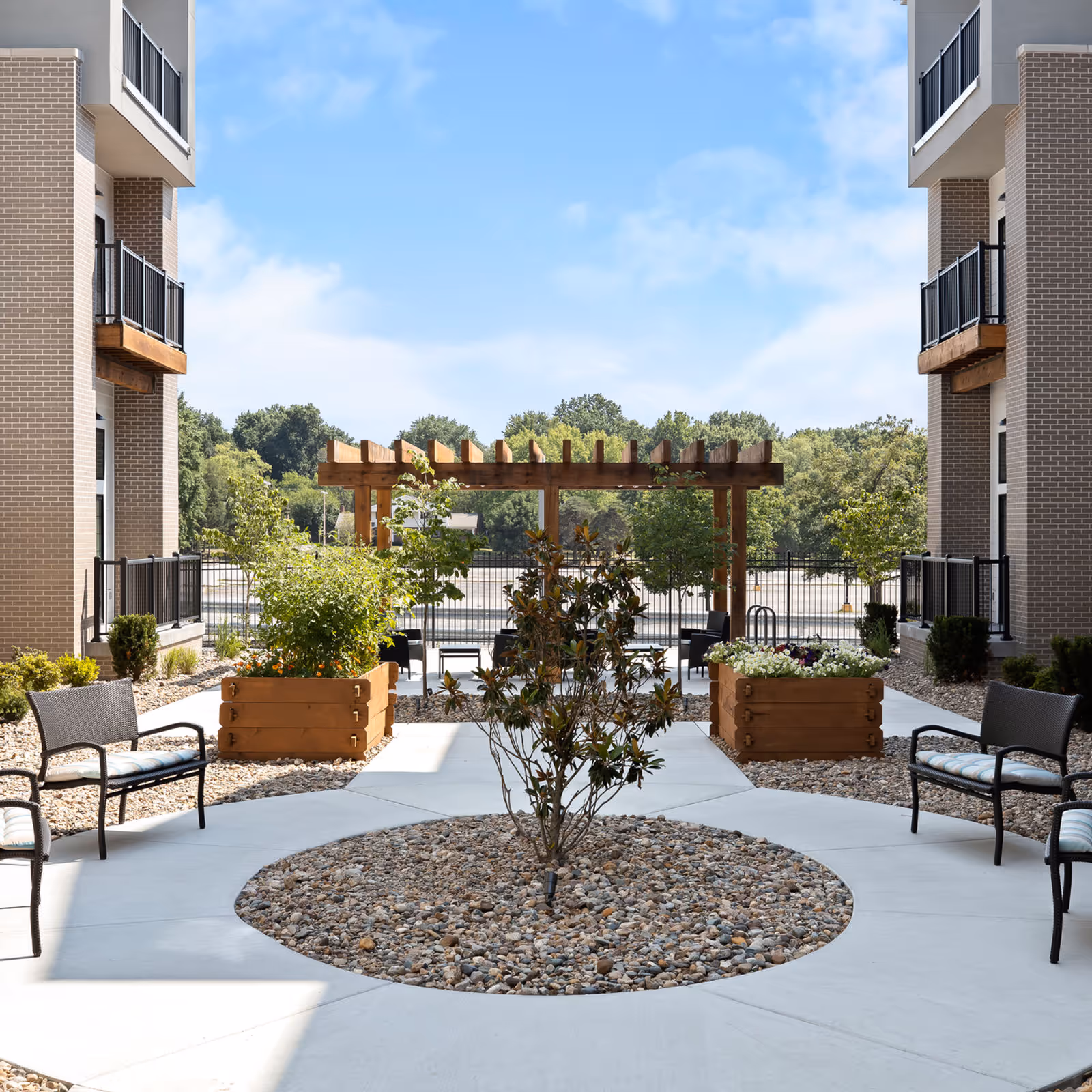 Outdoor courtyard area between two brick buildings with black metal balconies. The courtyard features a circular rock garden with a small tree in the center, wooden planter boxes with plants, black metal chairs with cushions, and a wooden pergola in the background under a blue sky with some clouds.
