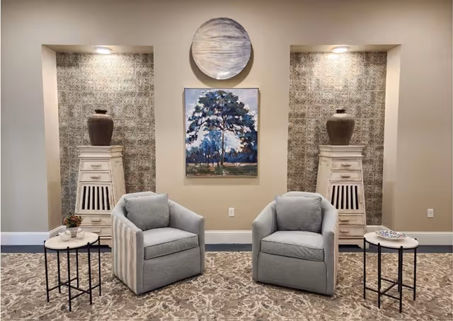 A cozy seating area with two gray upholstered armchairs facing forward, each accompanied by a small round side table. Behind the chairs are two tall white wooden cabinets with decorative vases on top, set into wall niches with patterned wallpaper and overhead lighting. A painting of a tree and a round decorative piece hang on the beige wall between the niches. The floor is covered with a patterned carpet.