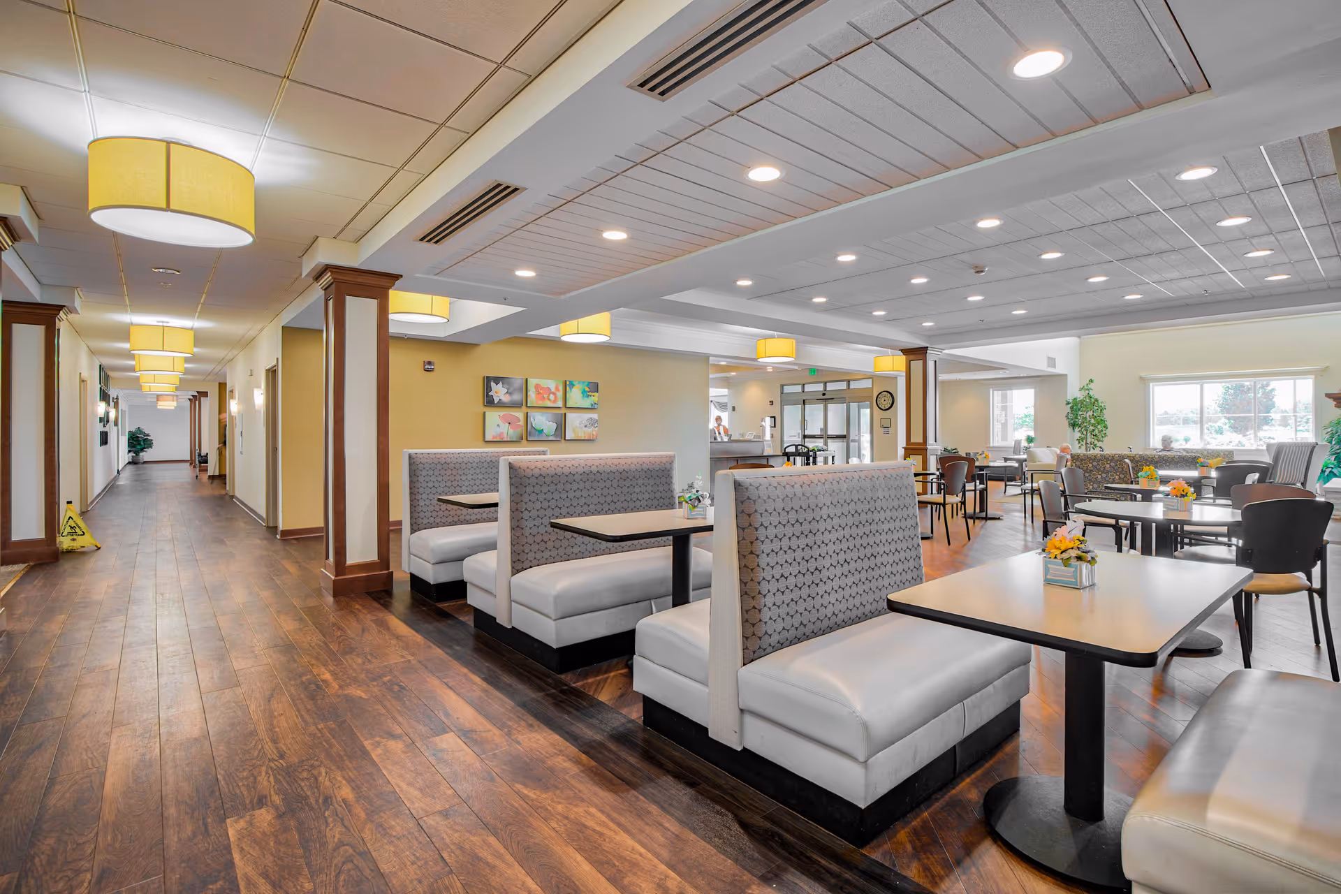 Interior view of a senior living facility dining area with booth seating and tables with chairs. The space has wooden flooring, yellow walls, ceiling lights, and decorative artwork on the walls. A long hallway is visible on the left side, and large windows provide natural light on the right side.