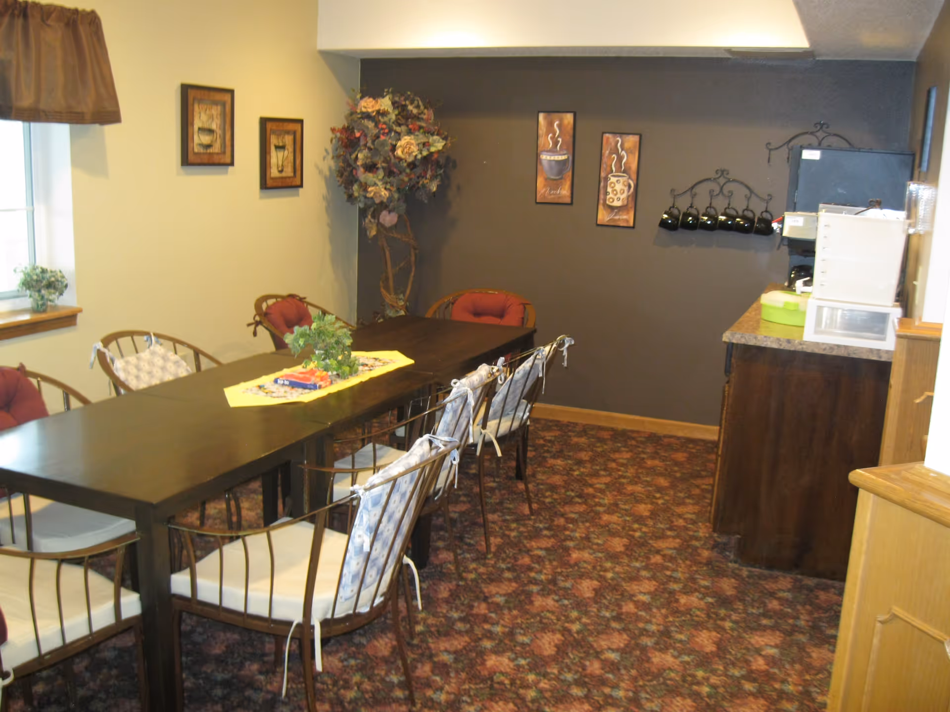 A cozy dining room with a long dark wooden table surrounded by metal chairs with cushions. The room has a patterned carpet, a window with a brown valance, and framed coffee-themed artwork on the walls. There is a small counter area with a coffee machine and cups hanging on a rack on the wall.