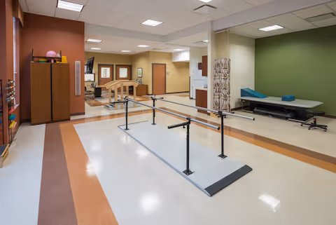 A spacious physical therapy or rehabilitation room with parallel bars for walking exercises in the center. The room has a brown and beige color scheme with a green accent wall. There is a treatment table with blue cushions on the right side, a wooden cabinet on the left, and various therapy equipment. The ceiling has recessed lighting and the floor is a mix of white and brown tiles.