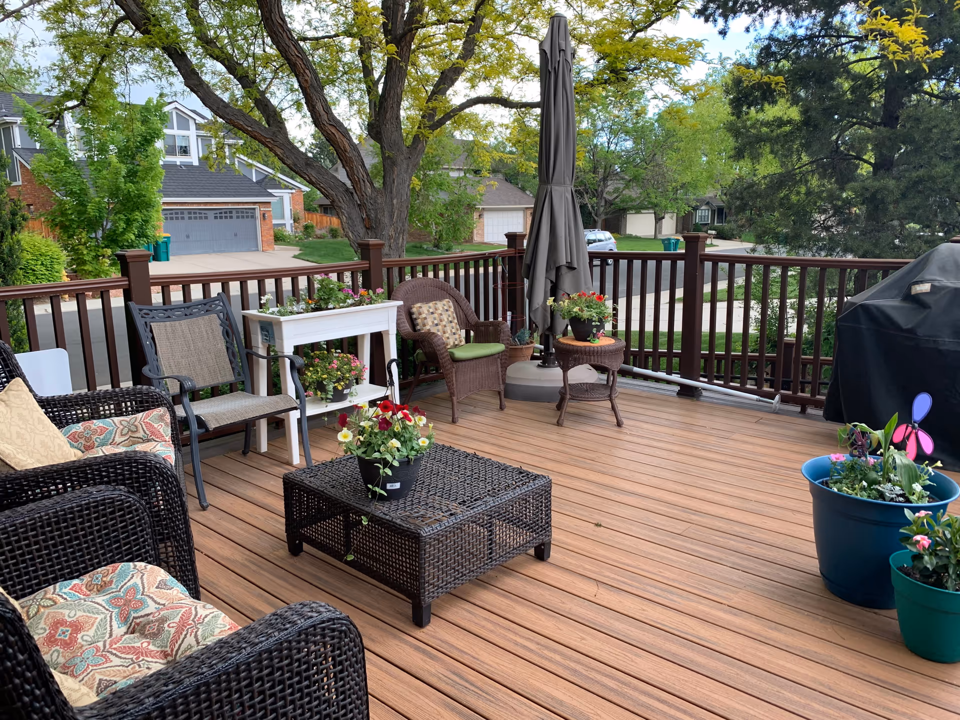 A wooden outdoor deck with wicker furniture including chairs and a coffee table, several potted plants with flowers, a closed patio umbrella, and a barbecue grill. The deck overlooks a suburban street with houses and trees in the background.