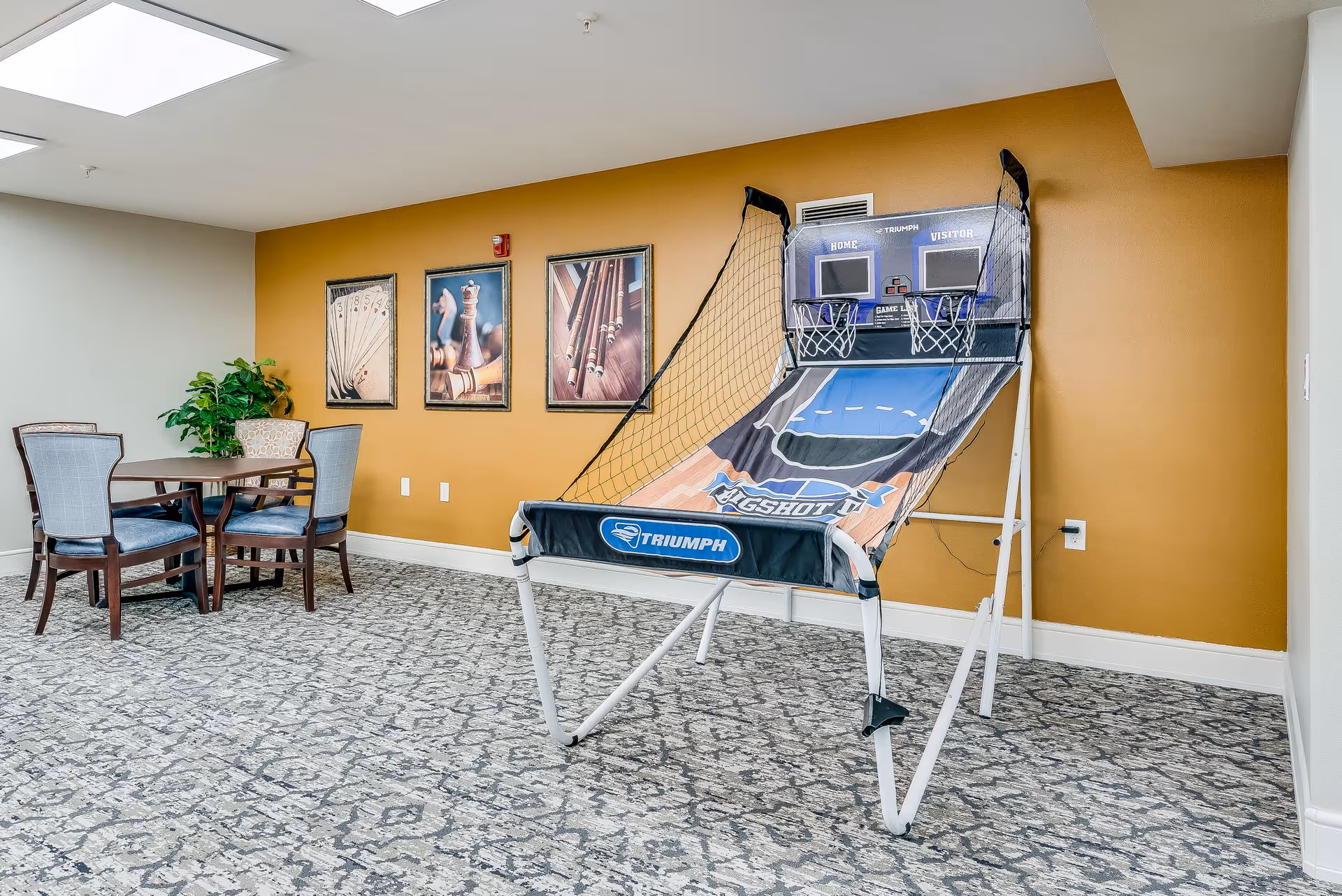 Indoor recreational area with a basketball arcade game and a small table with four chairs. The room has a patterned carpet, mustard yellow accent wall with three framed pictures, and a potted plant on the table.