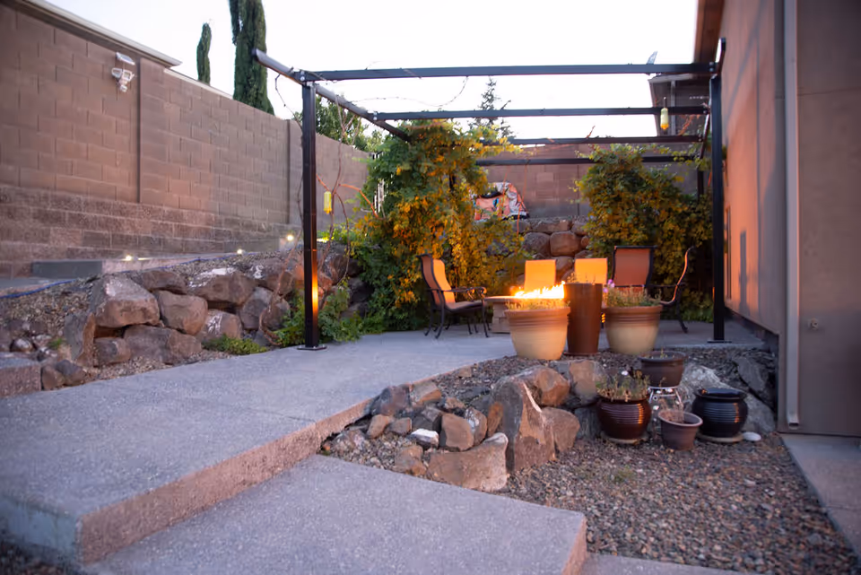 Patio with a pergola, chairs around a lit fire pit, potted plants, and rock landscaping beside a house and block wall.