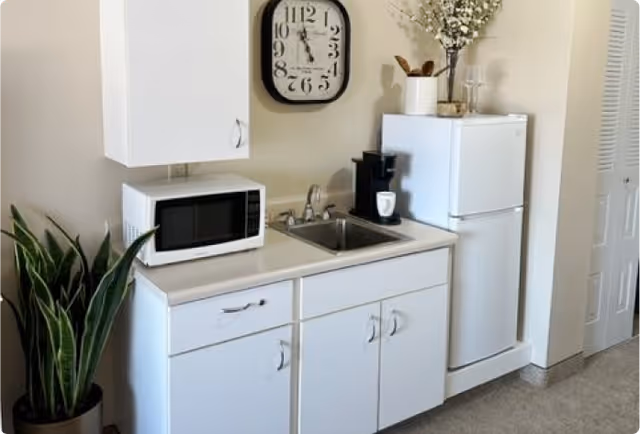 Small kitchenette with white cabinets, a sink, microwave, mini refrigerator, coffee maker, wall clock, and a potted plant.
