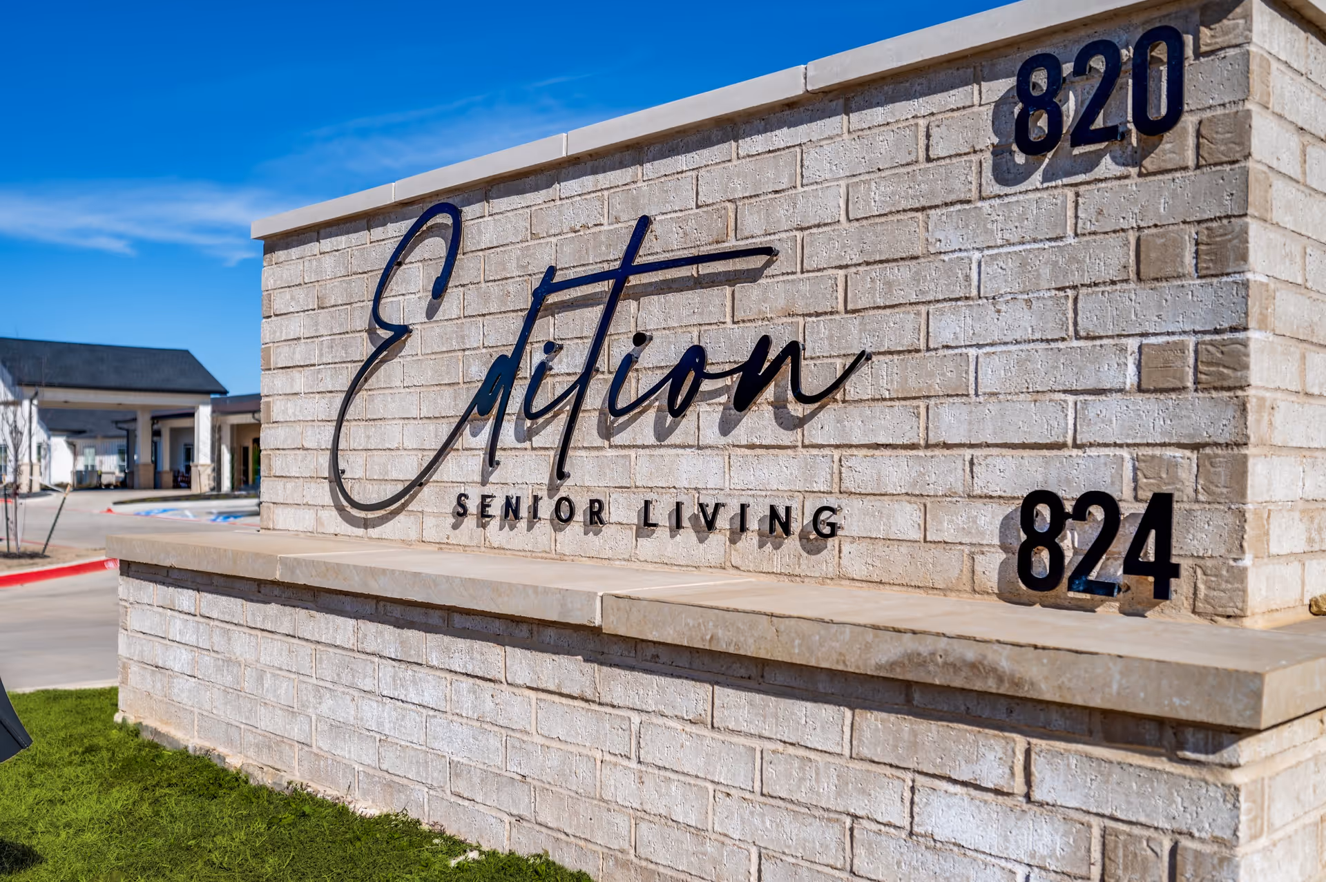 A brick sign for Edition Senior Living with the numbers 820 and 824 on the right side, set against a blue sky with part of the senior living facility building visible in the background.