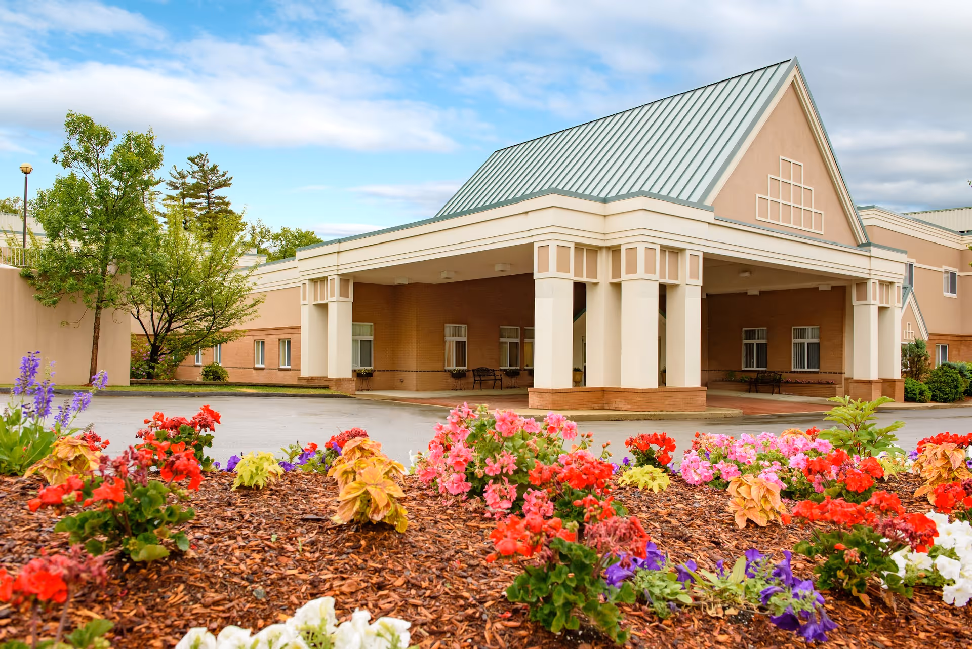 Entrance of a senior living facility with a covered drop-off area, beige walls, and a green metal roof. In the foreground, there is a colorful flower bed with red, pink, purple, and white flowers. Trees and shrubs surround the building under a partly cloudy sky.