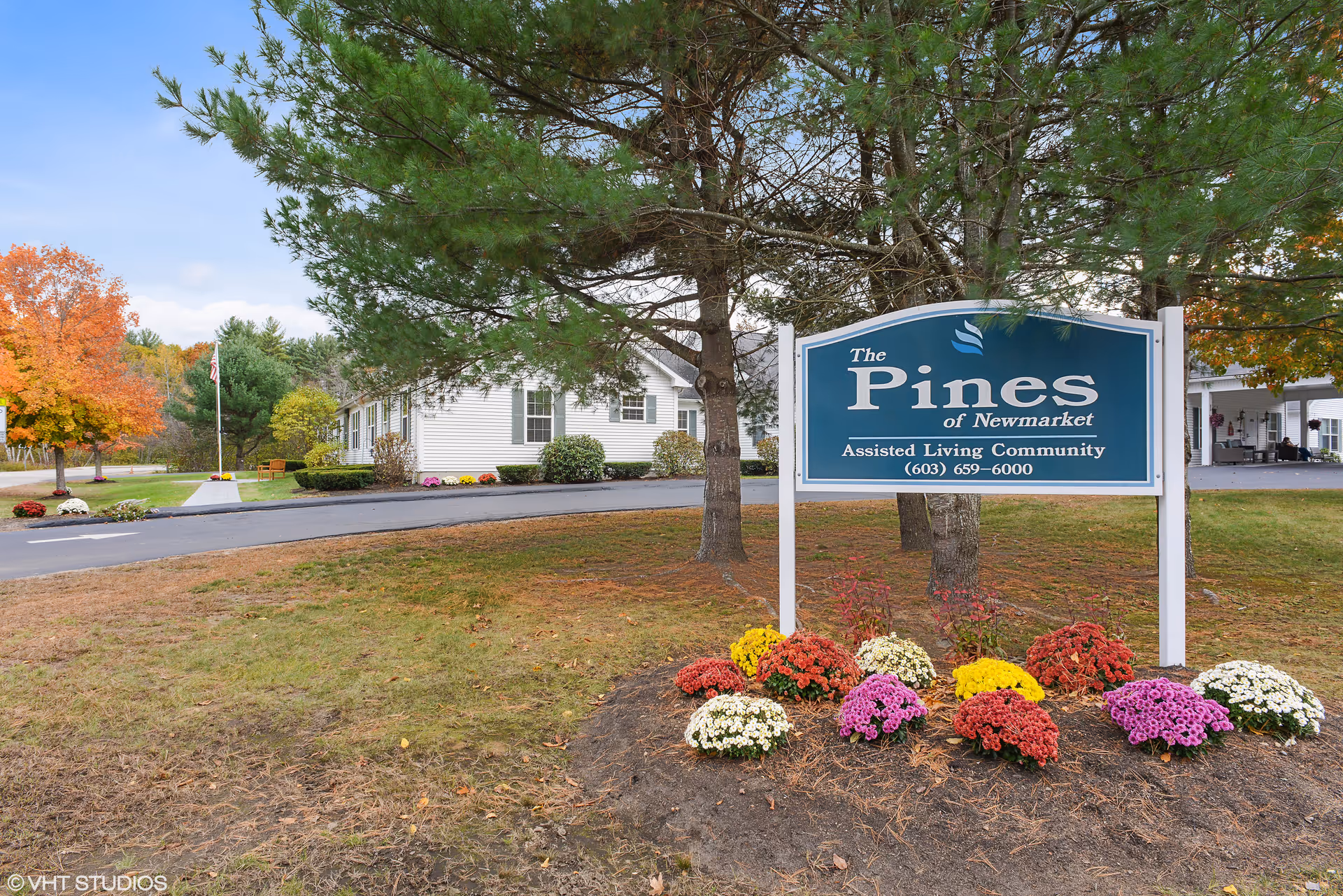 Outdoor view of The Pines of Newmarket assisted living community sign surrounded by colorful flowers and trees with autumn foliage, with a white building and driveway in the background.