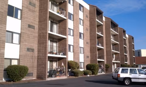 Multi-story brick senior living building with balconies and a parked SUV in a paved lot