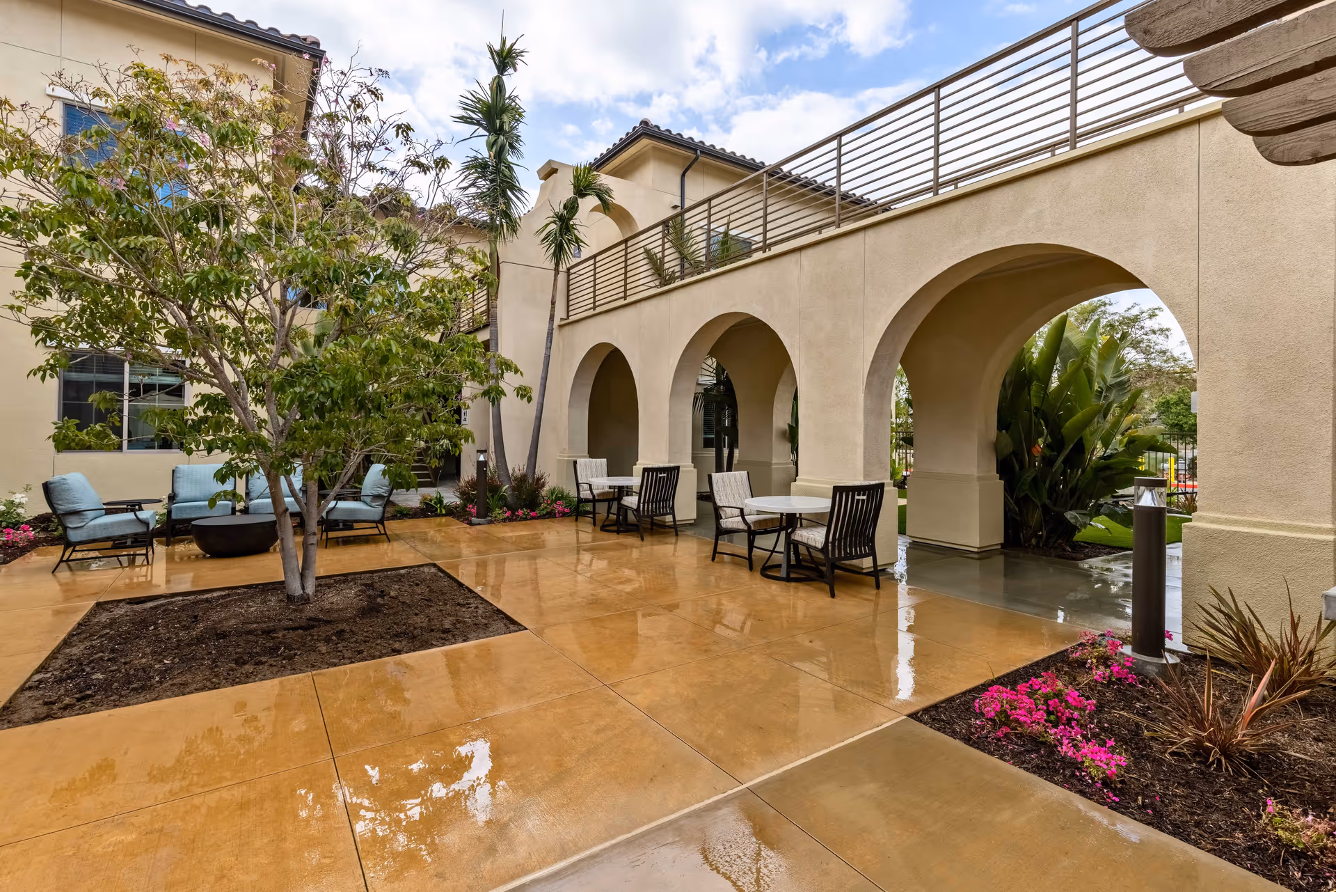 Outdoor courtyard area with wet tiled flooring, a tree planted in a square soil bed, several seating areas with chairs and tables, and a building with beige stucco walls and arched openings. There are plants and flowers around the edges and a partly cloudy sky above.