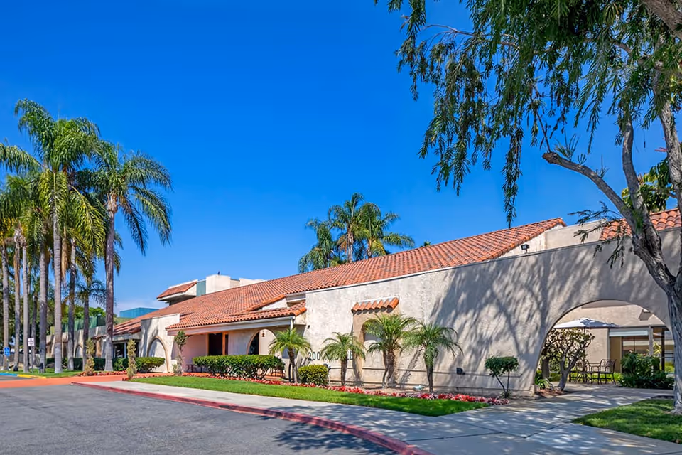 Exterior view of a senior living facility building with a red tile roof, beige stucco walls, and arched entryways. The building is surrounded by palm trees, green shrubs, and a well-maintained lawn under a clear blue sky.