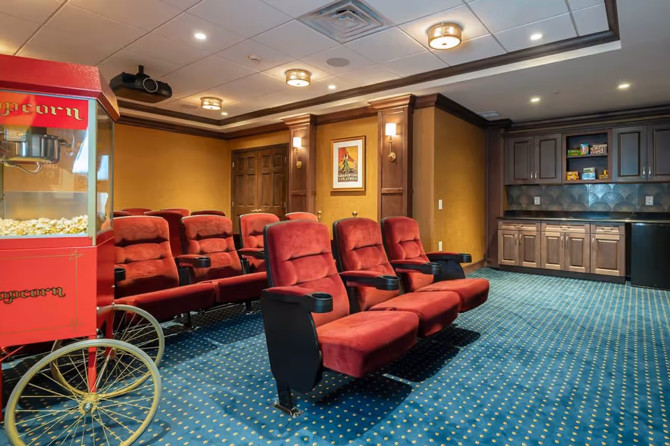 A cozy home theater room with two rows of red cushioned reclining seats, a red popcorn machine on wheels filled with popcorn, blue carpet with yellow dots, wooden cabinets with a black countertop, and warm yellow walls with framed artwork and wall sconces.