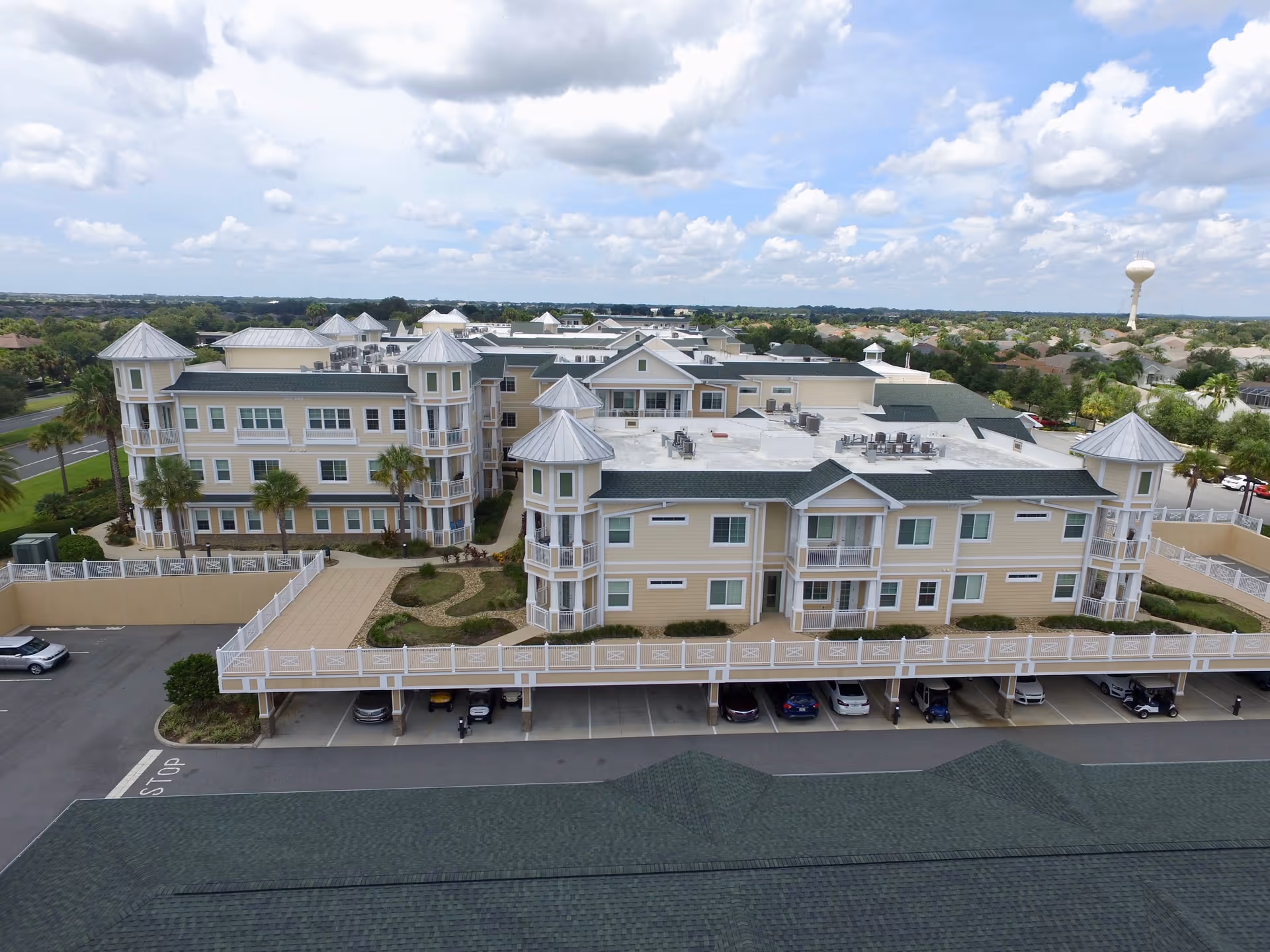 Aerial view of a large senior living facility with multiple beige buildings featuring green roofs and turret-style architectural elements. The buildings are surrounded by landscaped areas with palm trees and a parking area with several cars and golf carts. The sky is partly cloudy.